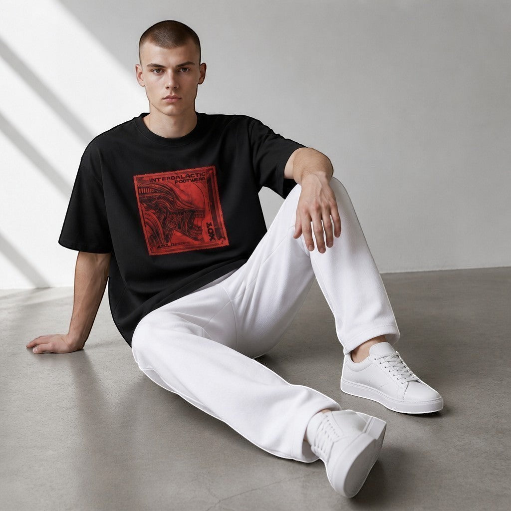 A model wearing a black vvicked oversized cotton t-shirt with a red alien stamp graphic, paired with white trousers.